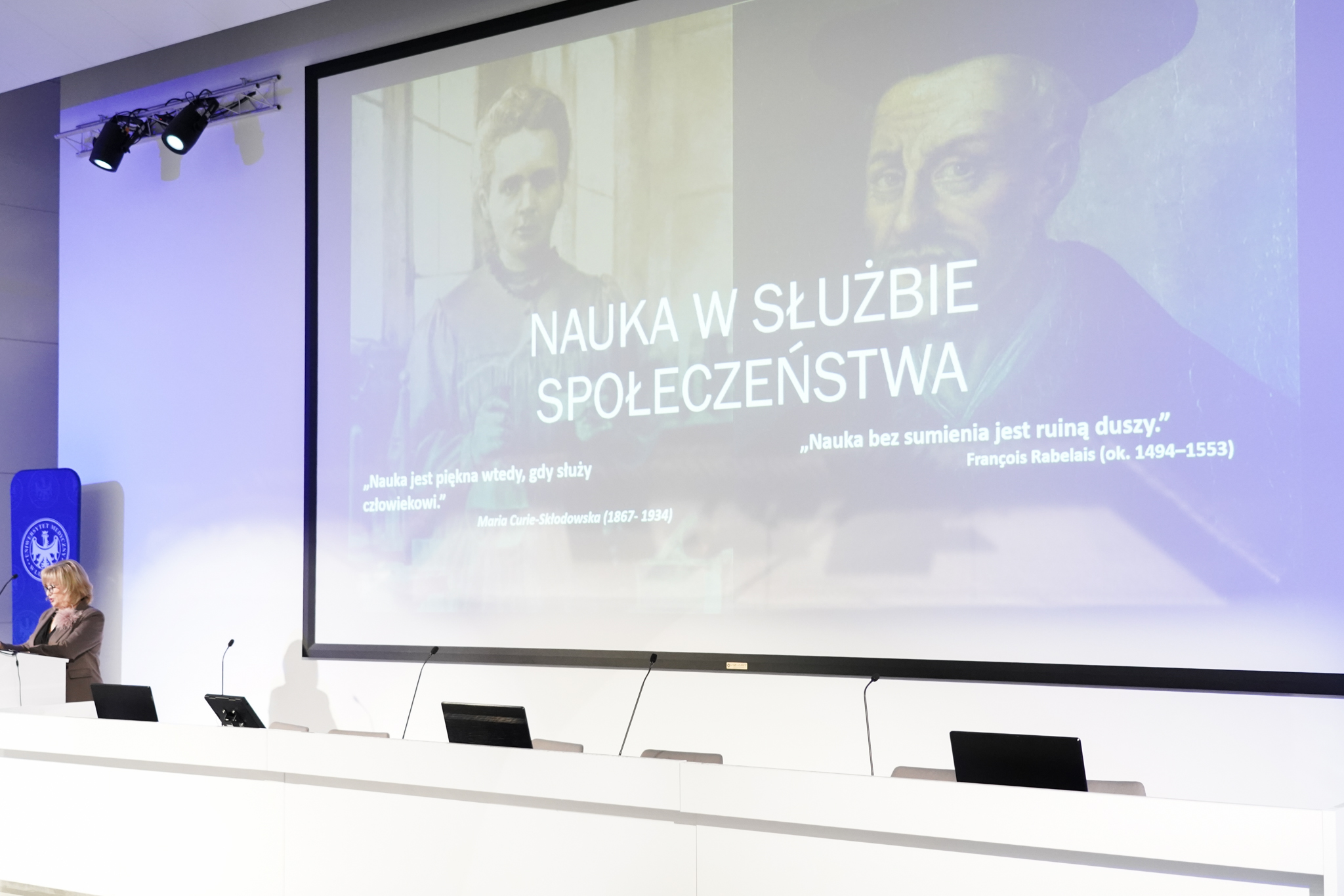 Na zdjęciu widać jasną salę z dużym ekranem zajmującym prawie całą środkową i prawą część fotografii. Na ekranie wyświetlony jest slajd z hasłem nauka w służbie społeczeństwa oraz dwoma cytatami, a w tle widać dwa portrety przedstawiające osoby z dawnych czas&oacute;w. Po lewej stronie slajdu, a także po lewej stronie zdjęcia, stoi kobieta przy białej m&oacute;wnicy, pochylona lekko nad komputerem przenośnym. Przed nią rozciąga się długi biały st&oacute;ł z kilkoma mikrofonami i zamkniętymi laptopami ustawionymi w r&oacute;wnych odstępach. W tle po lewej stronie stoi niebieski stojak z logo instytucji. Całość oświetlona jest jasnym, r&oacute;wnym światłem, kt&oacute;re podkreśla nowoczesny wygląd pomieszczenia.